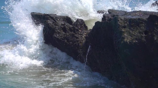 SLOW MOTION, CLOSE UP: Big foamy ocean waves crashing into black volcanic rock splashing waterdrops. Turbulent whitewater sea hitting against reef spraying water. Powerful tidal wave sparkling in sun