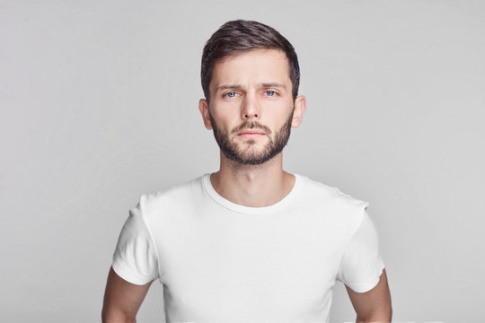 Close up portrait of good-looking serious bearded Caucasian man with blue beautiful eyes wearing white casual t-shirt posing isolated on gray studio wall with copy space for your promotional content.