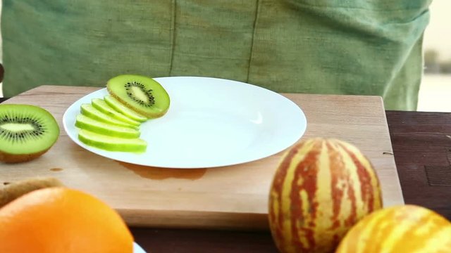 A woman puts pieces of kiwi, which is on a cutting board, on a plate.