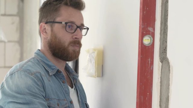 Male architect with smartphone checking wall with level at new apartment 
