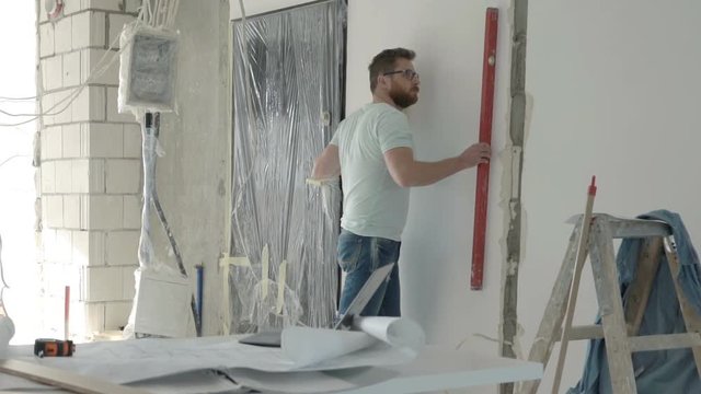 Male architect with blueprints checking wall with level at new apartment 
