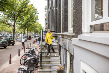 Young woman in yellow raincoat taking down a bicycle on the stairs near the beautiful residential house in Amsterdam