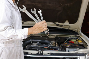 Portrait of professional young mechanic man in white uniform holding wrenches against car in open hood at the repair garage.