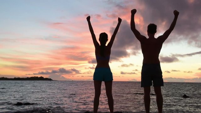 Rear view of young couple with arms raised by sea. They are in swimwear cheering at beach during sunset. They are representing concepts of success and accomplishment.