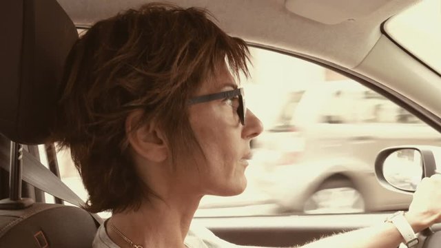 Woman with eyeglasses driving car with hands on steering wheel. Interior close up, side view, traffic on street. Vintage filter, toned image, slow motion.