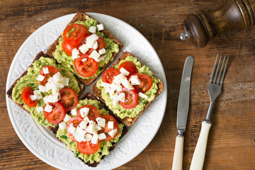 Toasts with mashed avocado, tomatoes and crumbled feta cheese on white plate on wooden table. Top view. Healthy eating, healthy snack concept