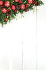 Christmas background border with red and gold bauble decorations, holly, mistletoe, ivy, juniper fir and pine cones on rustic white wood background.