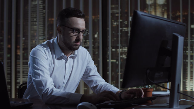 Man sitting and thinking while typing on computer in office at night.