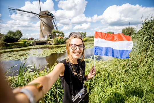 Young woman tourist making selfie photo with dutch flag standing on the beautiful landscape background with old windmills in Netherlands