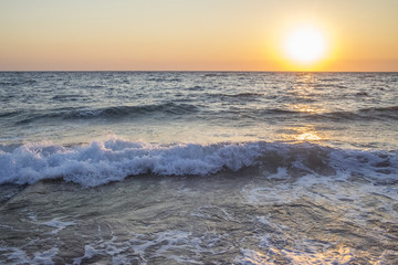Panorama del sole che tramonta a mare. L'acqua è mossa e le onde a contatto con la sabbia formano la schiuma. Il mare è agitato e il cielo è arancione per via del crepuscolo.