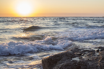 Obraz premium Panorama del sole che tramonta a mare. L'acqua è mossa e le onde a contatto con la sabbia formano la schiuma. Il mare è agitato e il cielo è arancione per via del crepuscolo.