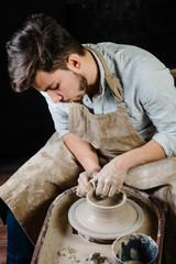 pottery, workshop, ceramics art concept - man working on potter's wheel with raw clay with hands, a male brunette sculpt a utensils near wooden table with tools, master in apron and a shirt