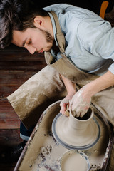 pottery, workshop, ceramics art concept - man working on potter's wheel with raw clay with hands, a male brunette sculpt a utensils near wooden table with tools, master in apron and a shirt