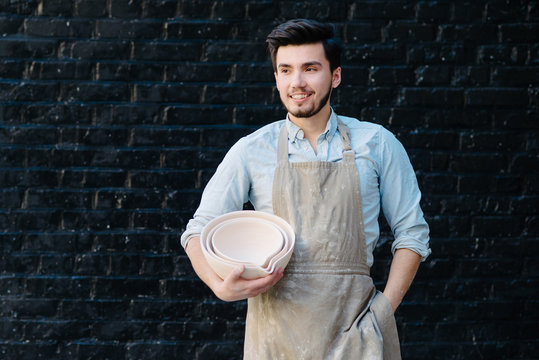 potter, utensils, ceramics art concept - smiling male ceramist in the apron shows samples of clay products, craftsman holding new finished bowls, happy master on background of dark gray brick wall