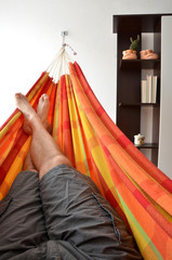 Legs of man lying down in bright hammock attached on house wall next to book-shelf
