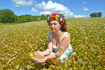 The beautiful woman of average years with a wreath on the head holds a bowl with buckwheat in the field of the blossoming buckwheat