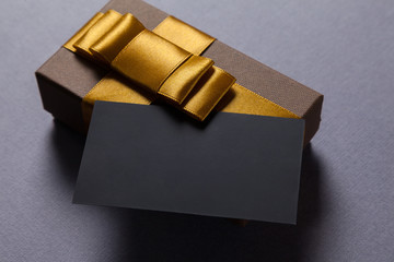 Blank black business card on gray table. Little gift box brown color with golden ribbon. Place for...