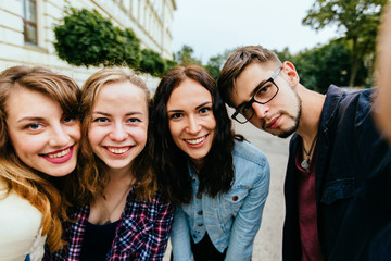 Focus on four positive student friends taking selfies and posing arm in arm during univercity park background .They are wearing summer clothes, guy in glasses.