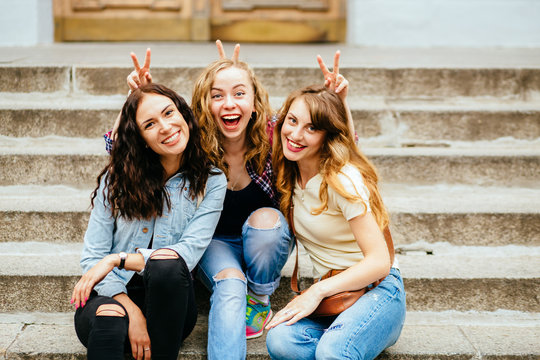 Three female laughing students makes bunny ears while sitting outdoor on university steps - education, school, teamwork and people concept.