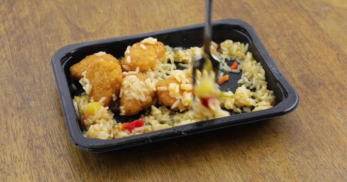 Sweet and sour chicken TV dinner in a black plastic microwavable tray being eaten with a fork on an old wood table top..