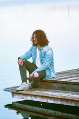 Porträt eines gutaussehenden jungen  Mannes mit langen Haaren sitzend auf einem Steg am See und genießt die Ruhe.  