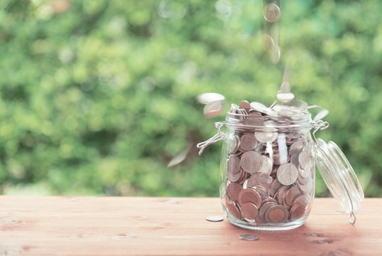 Money coins falling in glass jar on old  wooden table with green nature background,saving money concept