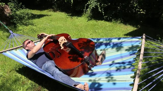 Mann liegt im Sommer entspannt in Hängematte und spielt auf dem Kontrabass