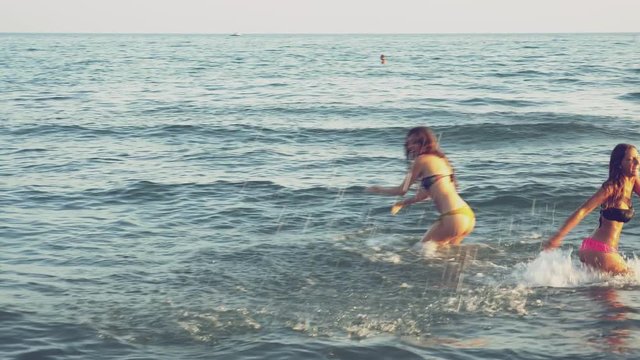 Two happy female friends in bikini splashing in the ocean having fun at sunset 4K
