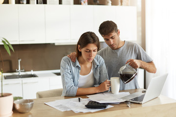 Fototapeta premium Domestic finances, home accounting, taxes, money and financial problems concept. Worried young American man and woman managing family budget together at home and having morning coffee in kitchen
