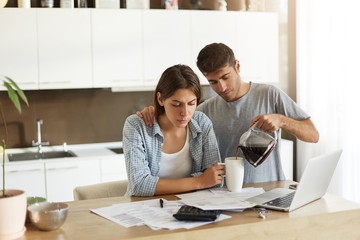 Obraz premium Picture of young male and female doing paperwork together at home: serious wife sitting at dining table with papers and laptop computer, calculating bills while her husband serving coffee to her
