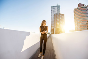 Lifestyle portrait of a stylish woman walking on the modern bridge with skyscrapers on the background during the morning in Amsterdam city