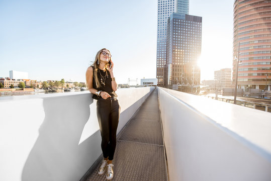 Lifestyle portrait of a stylish woman standing with phone on the modern bridge with skyscrapers on the background during the morning in Amsterdam city