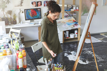 Creative female making strokes of brush on easel standing in her workshop, surrounded with different colorful oils. Talented painter drawing picture in art studio using watercolors and paint brush.