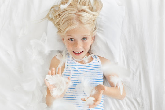 Happy childhood concept. Leisure, fun and relaxation. Top picture of adorable blonde freckled little girl preschooler looking at camera through flying feathers after pillow fighting in her room