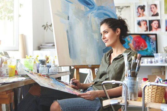 Thoughtful dreamy young European female artist bringing her creativity to life, sitting in her modern workshop interior with palette and painting knife. Hobby, job, occupation, art and craft concept