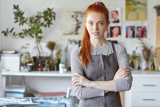Indoor portrait of charming confident young redhead crafts woman wearing grey apron standing in her workshop, keeping her hands crossed and smiling, ready for process of creation and hard work