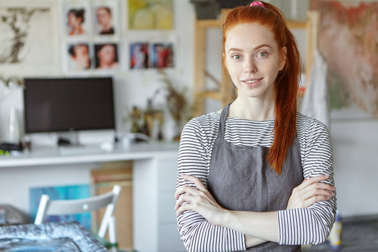 People, job, occupation, hobby and creativity concept. Picture of positive red haired young woman of creative profession wearing apron over striped top, posing in her studio with arms folded