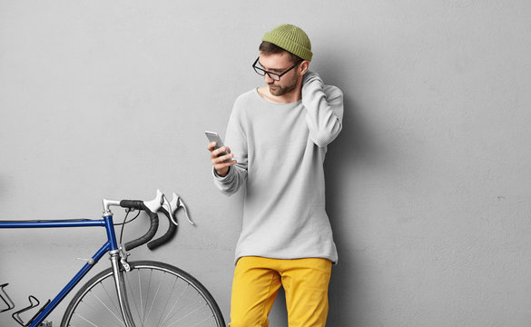 Unshaven Young European Guy Wearing Stylish Glasses And Hat Touching His Neck While Reading Important News Or Text Message On Generic Smart Phone, Standing At Grey Wall With Fixed Gear Bike