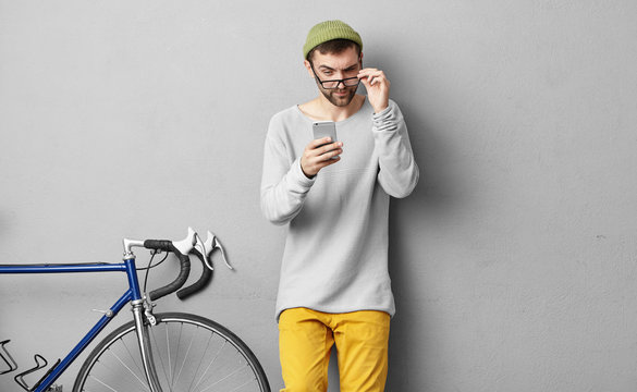 Picture of young bearded male wearing trendy hat and clothes holding eyeglasses while reading strange text message from unknown number, peering at screen, having suspicious or distrustful look
