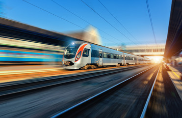 High speed train in motion at the railway station at sunset in Europe. Modern intercity train on the railway platform with motion blur effect. Industrial scene with moving passenger train on railroad
