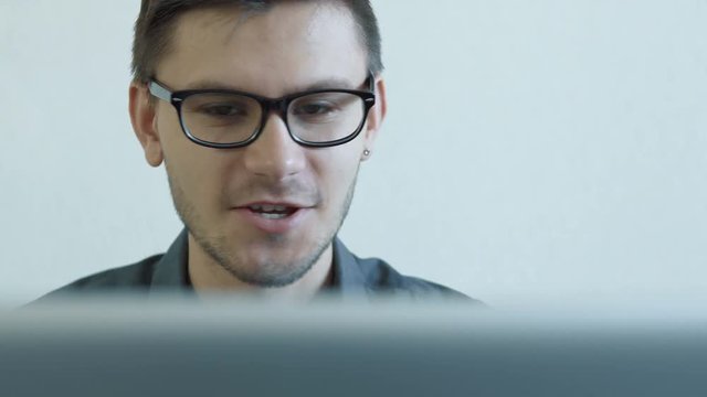 Close-up portrait of a young man wearing glasses sitting in his office in front of a monitor - having a video chat. People stock footage shot. 