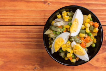 Flat lay of vegetable salad with boiled eggs, corn, carrot and potatoes above wooden table