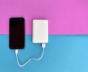 Closeup A black smartphone charging with white powerbank on pink & blue background. Black and white gadgets on colorful background. Top view with Copy space. Selective focus.