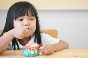 Asian children cute or kid girl happy to delicious eating dessert or milk ice cream in colorful ceramic cup on the wood table in restaurant or cafe for background with space
