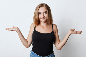 Beautiful female model with natural skin, blonde straight hair, wearing black t-shirt, shrugging shoulders while having uncertainty what to do. Attractive young female modeling in white studio