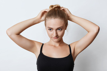 Fototapeta premium Horizontal portrait of attractive female with blonde hair knot, looking pensively down with her blue eyes while trying to make bun, thinking over where to go for walk with friends. Beauty concept