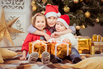 Fototapeta na wymiar Three children in hats of Santa Claus sitting with gifts and rejoice