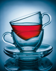 Two cups of transparent glass one inside the other on two glass plates at the top of the Cup poured tea, the whole composition is on a blue background and reflected on the surface
