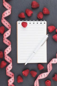 Red raspberries, tape measure, notebook and a pen on the black slate stone background