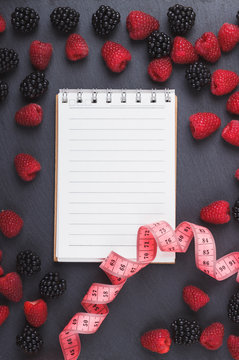 Red raspberries, tape measure, notebook and a pen on the black slate stone background